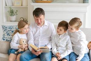 Caring caucasian father sitting on couch at home with young children, reading photo