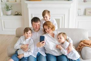 padres felices con tres hijos usando un teléfono inteligente en casa juntos sentados en un lugar acogedor foto