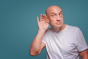 un hombre de fondo azul sosteniendo sus manos cerca de su oído, escuchando a escondidas