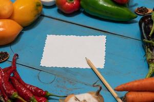 Vegetables are laid out around a sheet of paper and a pencil. Empty space for text. Vegetables, empty blank for recipe on a blue background. photo