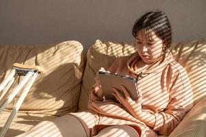 Girl with a cast on their leg, sitting on the couch and drawing on a graphics tablet. Bandaged leg cast and toes after a running injury accident.Teen girl in a plaster cast. photo