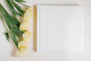 White family or wedding photo album with leather cover with empty space for text surrounded by yellow tulips on a light background. Family photo archive
