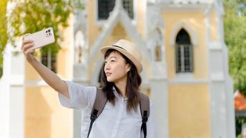 Young Asian woman traveler wearing white shirt and hat making picture on smartphone, happy female tourist posing for selfie using mobile phone camera photo
