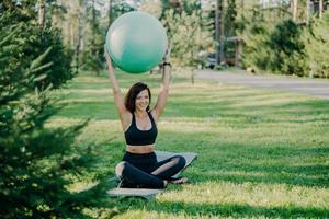 Horizontal shot of brunette fit woman in active wear sits in lotus pose on karemat holds fitness ball over head has gymanstic exercises outdoor during sunny day, smiles pleasantly, enjoys nature