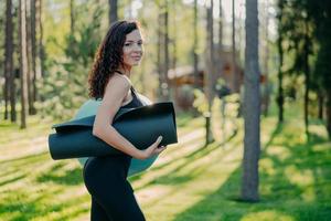 Sideways shot of sporty slim young woman dressed in black leggings, carries rolled up karemat under arm, poses against green grass and trees background, going to have exercises with fitness ball photo