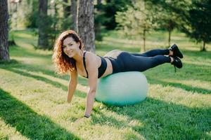 Sport, recreation, fitness and healthy lifestyle concept. Brunette sporty young European woman in active wear leans on fitness ball, makes physical exercises outdoor, enjoys favorite activity photo