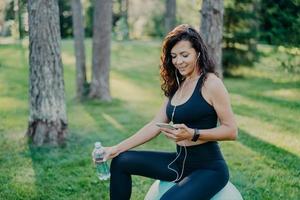 Pleased relaxed woman has rest after yoga exercises sits on fitness ball, listens music via modern mobile phone and earphones, holds bottle of fresh water, dressed in active wear, poses at park photo