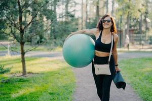 una foto al aire libre de una mujer morena feliz y delgada vestida con ropa activa gafas de sol de moda lleva equipo deportivo a pie disfruta del aire fresco y la hermosa naturaleza. hembra deportiva tiene fit ball y karemat