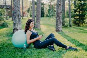 Relaxed sporty young female model takes break after aerobics exercises with fitness ball, sits on green grass, uses mobile phone, chats online, listens music in earphones, has athletic body. photo