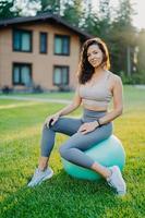 Vertical shot of beautiful brunette woman sits on fitness ball over green grass and house in background, dressed in active wear, has yoga or gymnastic training, being in good shape. Active lifestyle photo