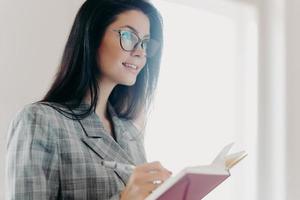 Serious thoughtful brunette European woman writes down text information in notepad, concentrated somewhere into distance, makes notes, wears formal clothes, transparent glasses, stands indoor photo