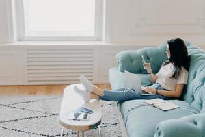 Sideways shot of relaxed female student uses modern technologies while doing homework, uses web sources for making project, sits on comfortable sofa against cozy interior, leans legs on desktop photo