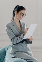 Vertical shot of successful female manager checks finance report, involved in working process, signs inportant contract, has happy expression, wears formal suit, poses in living room, works from home photo