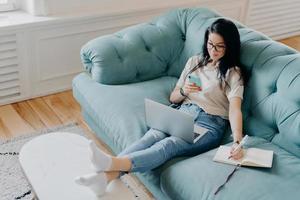 personas, aprendizaje a distancia, tecnologías, concepto de trabajo independiente. una mujer morena seria escribe información en el diario, sostiene un teléfono móvil, se concentra en un trabajo remoto, trabaja con una computadora portátil foto