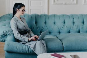 Content businesswoman works remotely in cozy home office, poses with laptop computer on sofa, wears formal elegant clothes, keyboards and searches necessary information in internet, sends emails photo