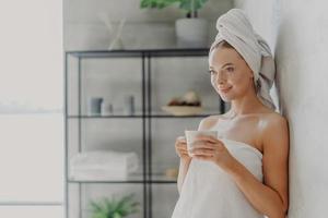 toma horizontal de una hermosa joven con piel sana envuelta en una toalla de baño, posa con una taza de té, concentrada en la distancia, posa cerca de la pared sobre el interior de la casa, disfruta de un ambiente acogedor foto