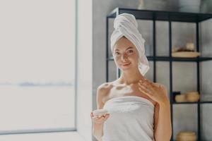 concepto de belleza e higiene. una joven atractiva y saludable tiene una piel suave y brillante, aplica crema corporal, sostiene un frasco de producto cosmético, se para envuelta en una toalla de baño, posa en un baño acogedor foto