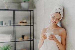 Pretty smiling woman with healthy skin wrapped in white towel, stands glad indoor, drinks coffee, has positive face expression, poses indoor. People, spa, pampering, skin care and wellness concept photo