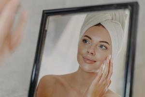 una joven europea desnuda toca la piel suave de la cara, se mira en el espejo del baño, usa una toalla blanca envuelta en la cabeza después de bañarse, posa los hombros desnudos. concepto de belleza y cuidado de la piel foto