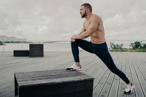 Motivated sportsman leans at knee and does sport exercises outdoor, warms up before jogging, poses near river and dressed in active wear. Male runner does stetching exercise, prepares for workout photo