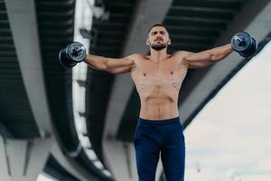 Horizontal shot of fit muscular bearded man works out with heavy weights trains muscles and raises arms being professional bodybuilder demonstrates determination stands powerful under bridge photo
