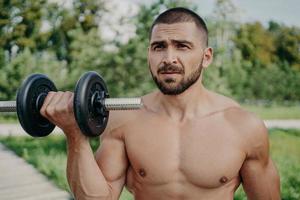 Serious sportsman poses with naked torso has muscular body raises dumbbell, poses outdoor, being in good physical shape, enjoys fresh air and nature. Strength, sporty lifestyle and bodybuilding photo