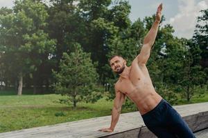 Strong young man stands in side plank on one arm, finds balance, poses at park near trees, practices yoga outdoor, leads active healthy lifestyle, has muscualr strong body. Motivated bodybuilder photo