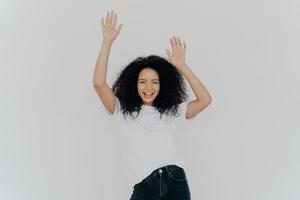 Half length shot of energetic carefree curly woman cheers with hands raised, wears white t shirt and jeans, has lucky day, perfect weekend, models against white background. Everybody put your hands up photo