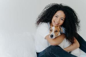 Photo of attractive young woman with Afro hircut, embraces with love dog, takes care of pet, smiles gently, wears casual clothing, isolated over white background, sit on bed, copy space for your promo