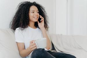 Sideways shot of positive ethnic brunette woman has telephone conversation, drinks hot beverage, holds white cup, sits on comfortable couch in living room, hears pleasant news, smiles happily photo