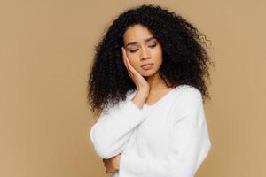 Melancholic sad lonely woman focused down with thoughtful expression, keeps hand on cheek, has calm expression, wears white sweater, has curly hair, isolated on brown studio wall. Depression photo