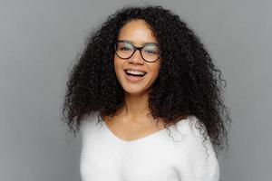 Candid shot of optimistic curly woman laughs positively, being in good mood, has healthy skin, looks through transparent glasses, wears white sweater, isolated over grey background. Happiness photo