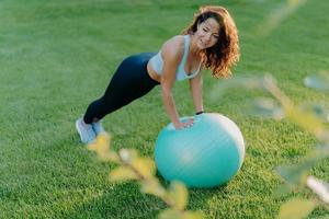 Pleased slim woman leans at fit ball, dressed in active wear, has gymnastic exercises on green lawn, enjoys sport and favorite activity, leads healthy lifestyle. People, aerobics, fitness concept