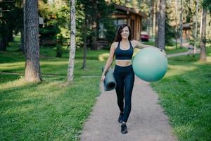 Horizontal shot of sportswoman wears cropped top and leggings, carries fitness ball and rolled up karemat, walk on road around trees and green grass, leads healthy lifestyle. Sport equipment photo