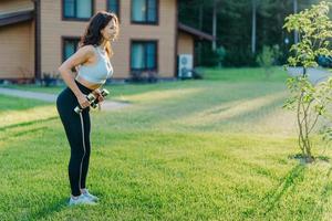 Shot of active sporty woman raises dumbbells and has workout session, works on biceps, dressed in top and leggings, poses on green grass near private house, has athletic body, leads active lifestyle photo