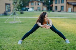 Confident athletic woman demonstrates her flexibility, stretches outdoors, does exercises early in morning on green grass near house leads active lifestyle dressed in active wear stays healthy photo