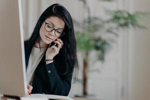 Stylish female entrepreneur in black suit, has phone conversation, makes notes in organizer looks assured and ambitious poses at desktop against office interior. Young woman manager talks via cellular photo