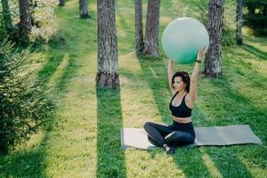 Fit sporty woman exercises with fitness ball sits on karemat in lotus pose dressed in cropped top and leggings, poses in forest during sunny day, breathes fresh air. Active lady practices yoga outside photo