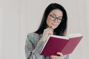 Serious brunette female management consultant uses notepad for planning organization, holds pen and writes down notes or list to do, wears formal clothing, has serious expression, looks in notebook photo