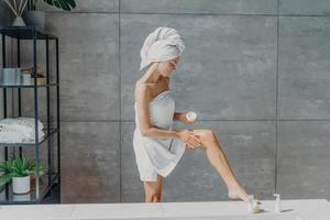 Indoor shot of slim female model applies leg cream stands wrapped in bath towel takes care of body and skin undergoes beauty treatments after taking shower poses in bathroom. Cosmetology concept photo