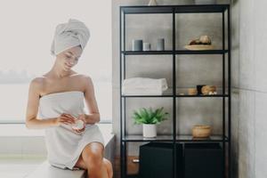 Daily routine, wellness and hygiene concept. Pleased young slim young European woman wrapped in bath towel applies body cream, has healthy skin, poses in cozy bathroom, takes care of herself. photo