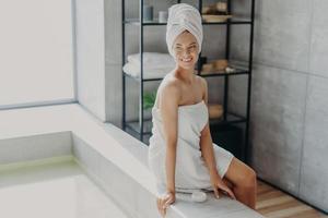 Beauty, grooming and hygiene concept. Relaxed beautiful European woman with healthy skin smiles and looks away, wrapped in bath towel, uses cosmetic cream, enjoys beauty routine, takes shower photo