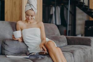 Beautiful young woman wrapped in bath towel, reads beauty magazine, drinks hot beverage, poses on comfortable couch in living room, applies face cream, has healthy smooth skin after taking shower photo