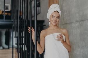 Healthy attractive European woman with pensive expression smiles gently, applies face cream on complexion, stands wrapped in towel, holds mug of drink, relaxes at home. Natural beauty concept photo