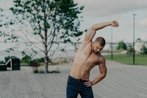 Active sportsman with naked torso and muscular body, does stretching exercises outdoor, shows good flexibility. Motivated athlete unshaven man warms up, prepares for workout, keeps muscles flexible. photo