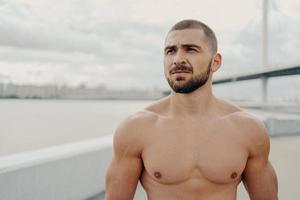 Horizontal shot of muscular man with naked torso thick beard looks somewhere into distance, relaxes outdoors, stands against river background, takes break after jogging, has athletic body shape photo