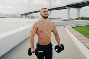 Muscular bodybuilder concentrated into distance has strong body strong muscles holds barbells and does exercises for biceps outdoor poses near river with bridge. Healthy lifestyle and sport concept