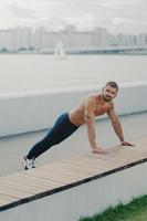 Outdoor shot of determined muscular man warms up before training stands in plank pose has naked torso muscular body enjoys exercising in open air wears active wear has workout to maintain vitality photo