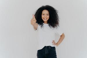 foto de una mujer feliz y rizada con el pelo rizado levanta el pulgar, da su aprobación, dice que suena bien, hace un gesto de apoyo, mantiene la otra mano en la cintura, vestida informalmente, aislada sobre fondo blanco