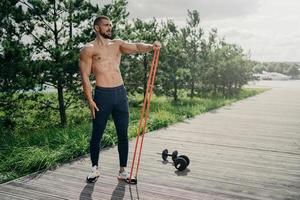 Full length shot of strong bearded sportsman flexes muscles with elastic resistance band, wears sport trousers and sneakers, stands outdoor, uses sport equipment for workout, performs exercises photo